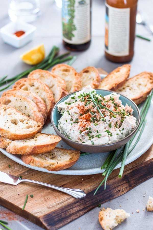 Smoked trout dip presented in a bowl sprinkled with chives and with toasted baguette chips.