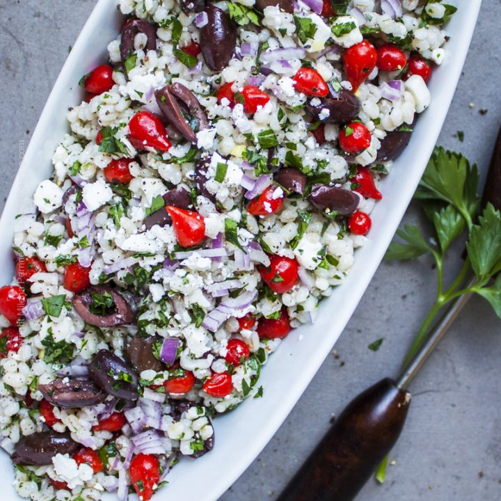 Barley salad with feta cheese, olives and sweety drops served in a white bowl.