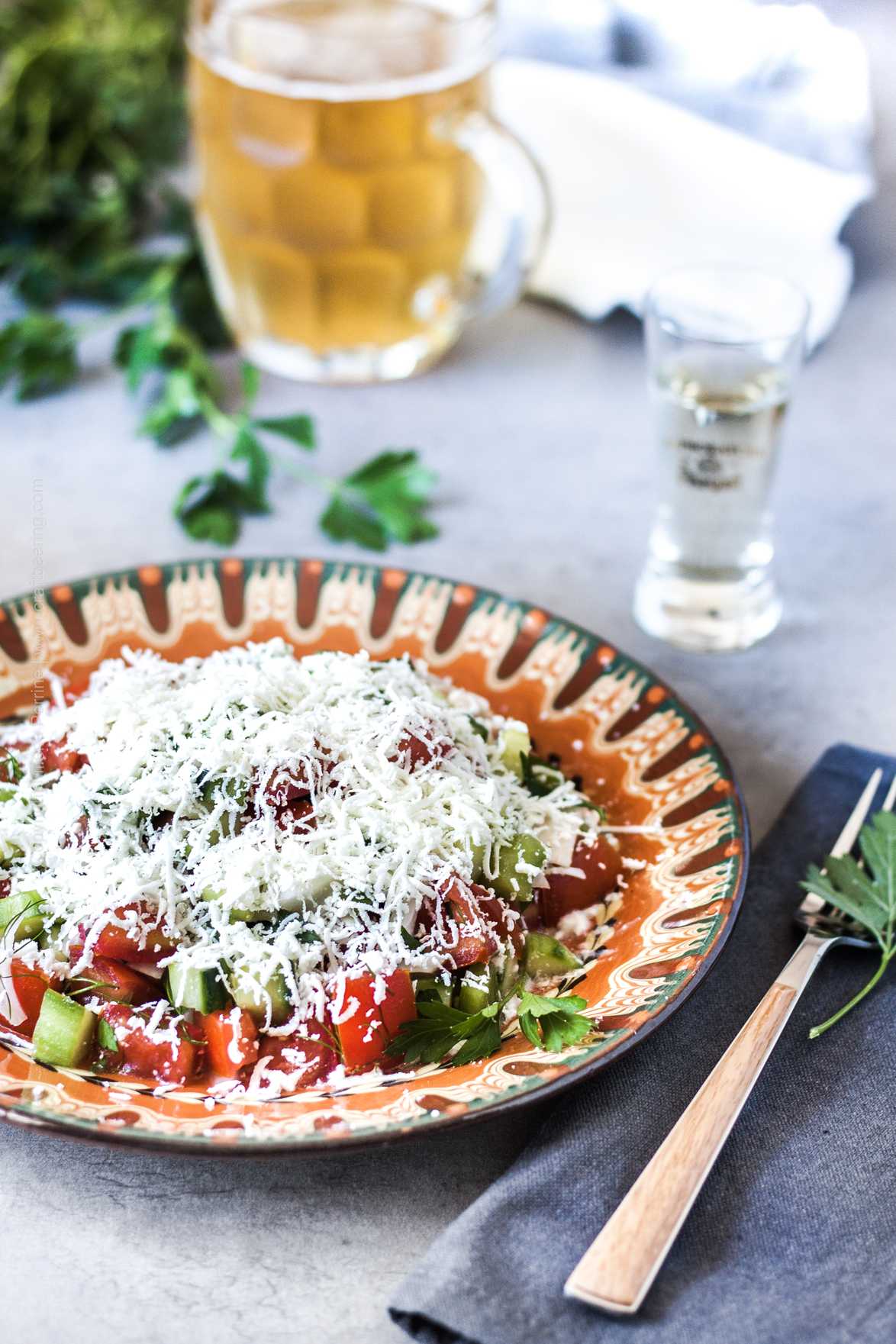 Shopska salata served in traditional Bulgarian plate along with a shot of rakiya. The feta is very finely grated on top of the cucumber, tomato, onion and pepper medley.