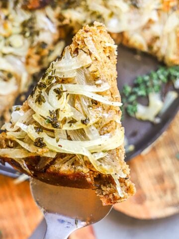 Onion tart slice is held up in focus over the backdrop of a plated onion pie.