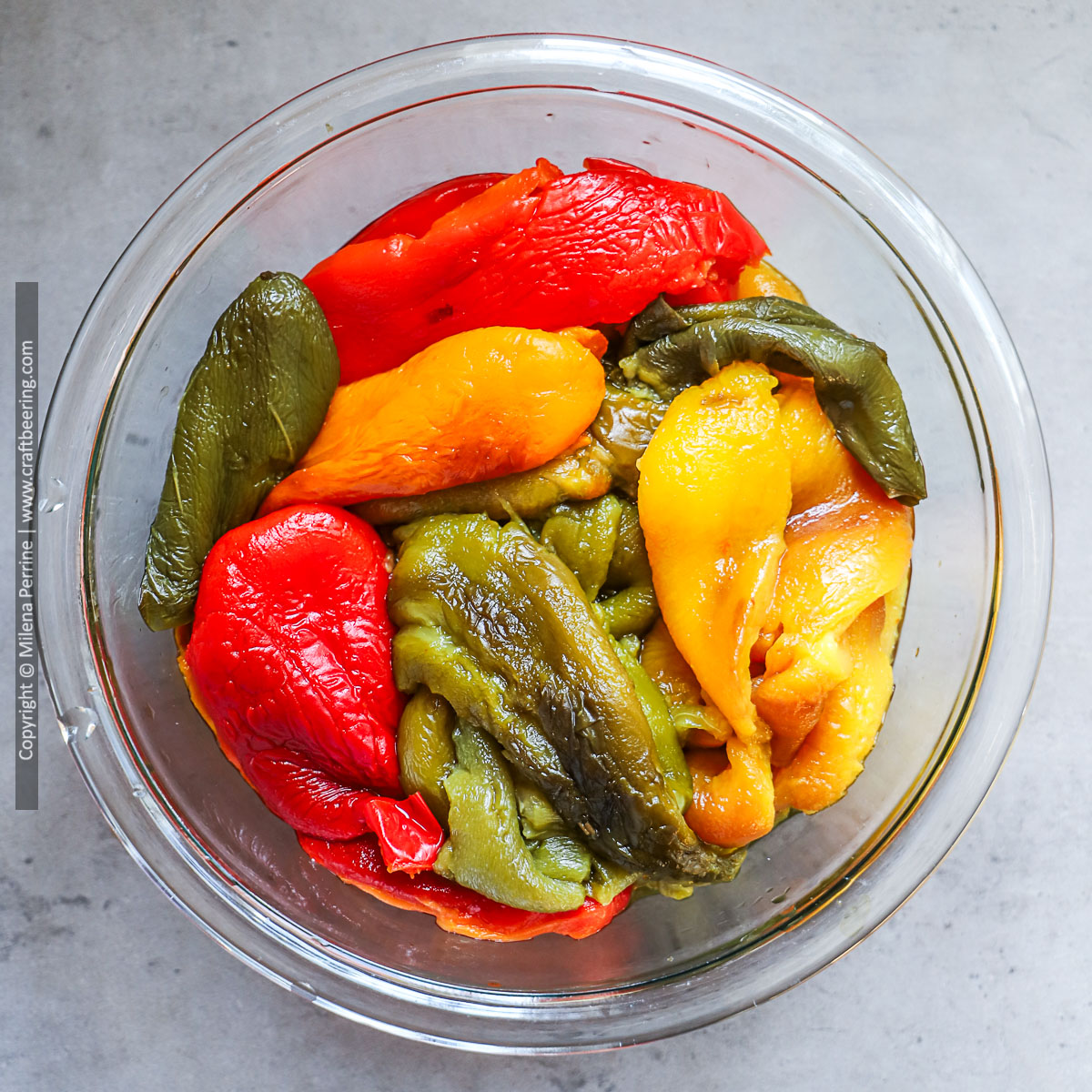 Roasted bell peppers of different colors are shown peeled and arranged in a clear glass bowl.
