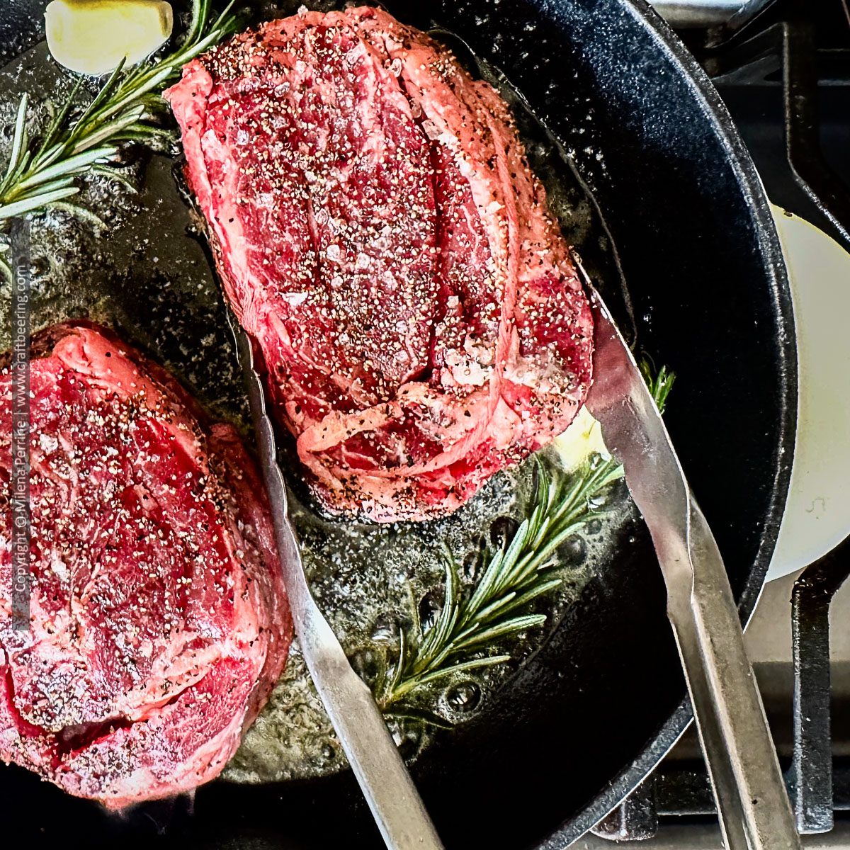 Chuck eye steak in in focus shown being seared in a cast iron skillet with rosemary and garlic.