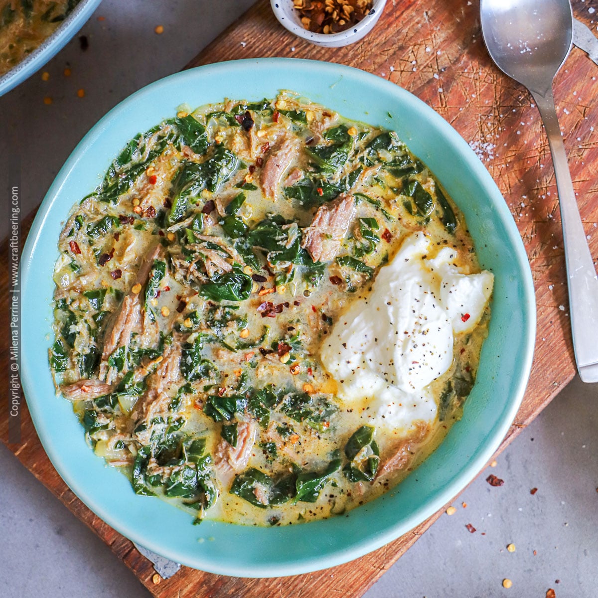 Lamb soup with spinach and egg is shown served in a light blue bowl, ganrished with yogurt, placed on top of a wooden board.