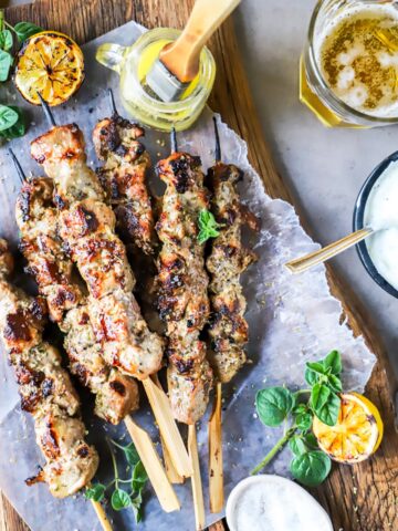 Pork souvlaki skewers are shown piled up on a wooden board with tzaziki sauce and lemons nearby.
