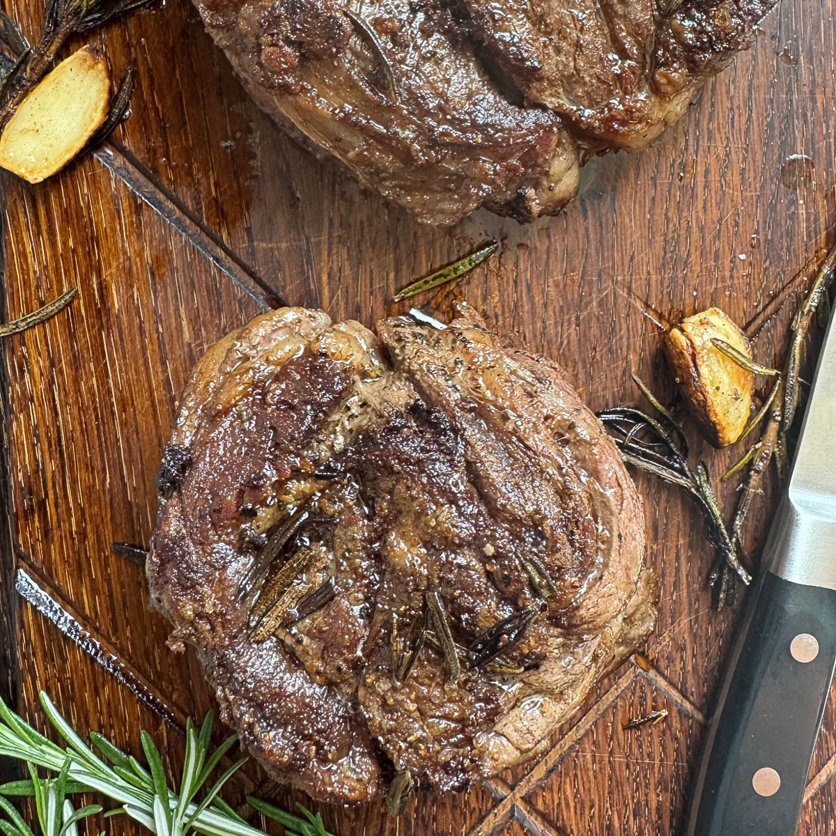 Two pan seared chuck eye steaks are shown over a wooden cutting board with garlic and rosemary