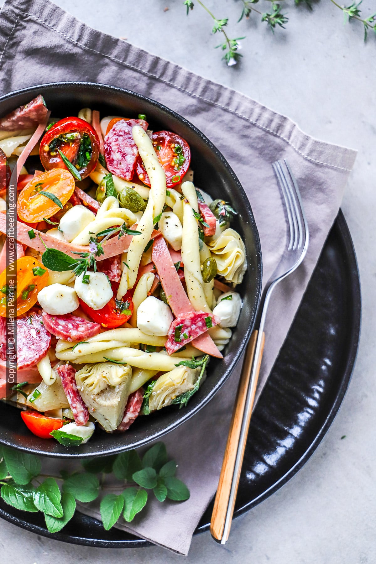 Antipasto pasta salad with colorful ingredients such as cherry tomatoes, capers, Italian salami and artichoke hearts is shown served in a black bowl.