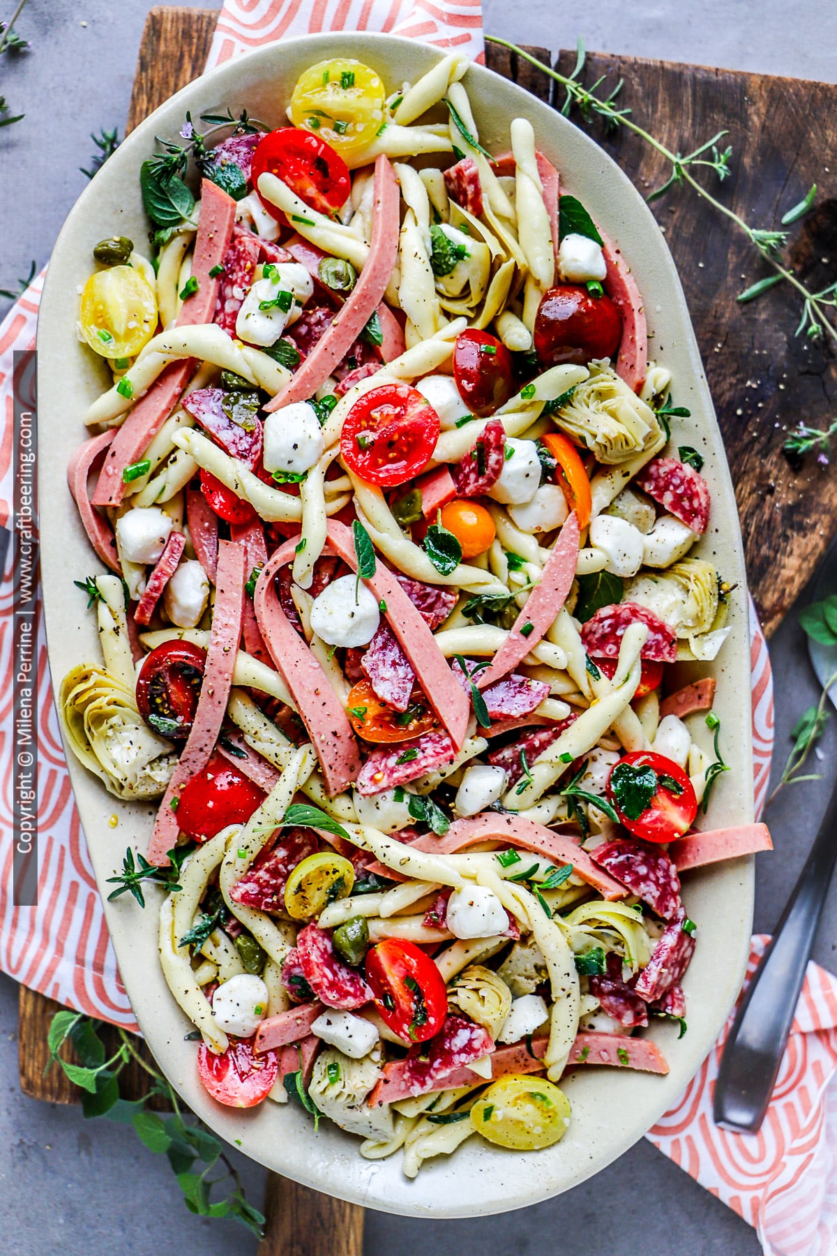 A colorful platter of antipasto pasta salad with cherry tomatoes, mozzarella pearls, artichoke hearts, salami, capers and more is shown viewed from above.