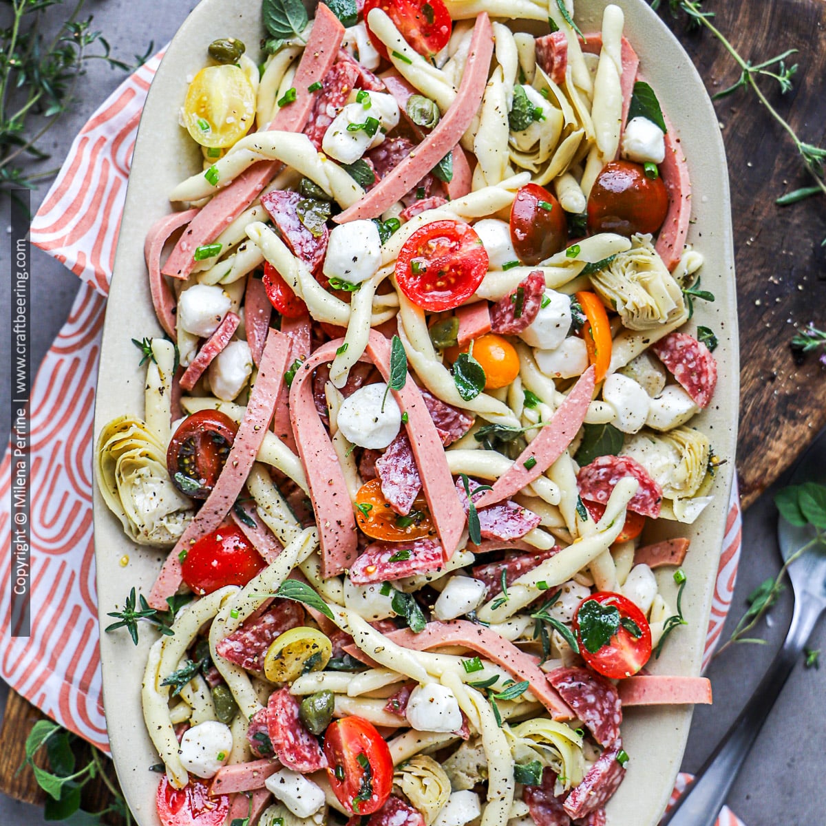 Antipasto pasta salad is shown from above with short grain pasta, salami, capers, artichokes, bologna strips, tomatoes, basil, Italian organo, mozzarella pearls and more.