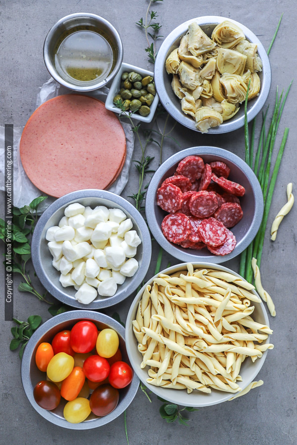 An sample arrangement of ingredients for pasta salad - al dente busiati noodles, hard salami, mozzarella pearls, fresh herbs, marinated artichokes , capers, Italian dressing.