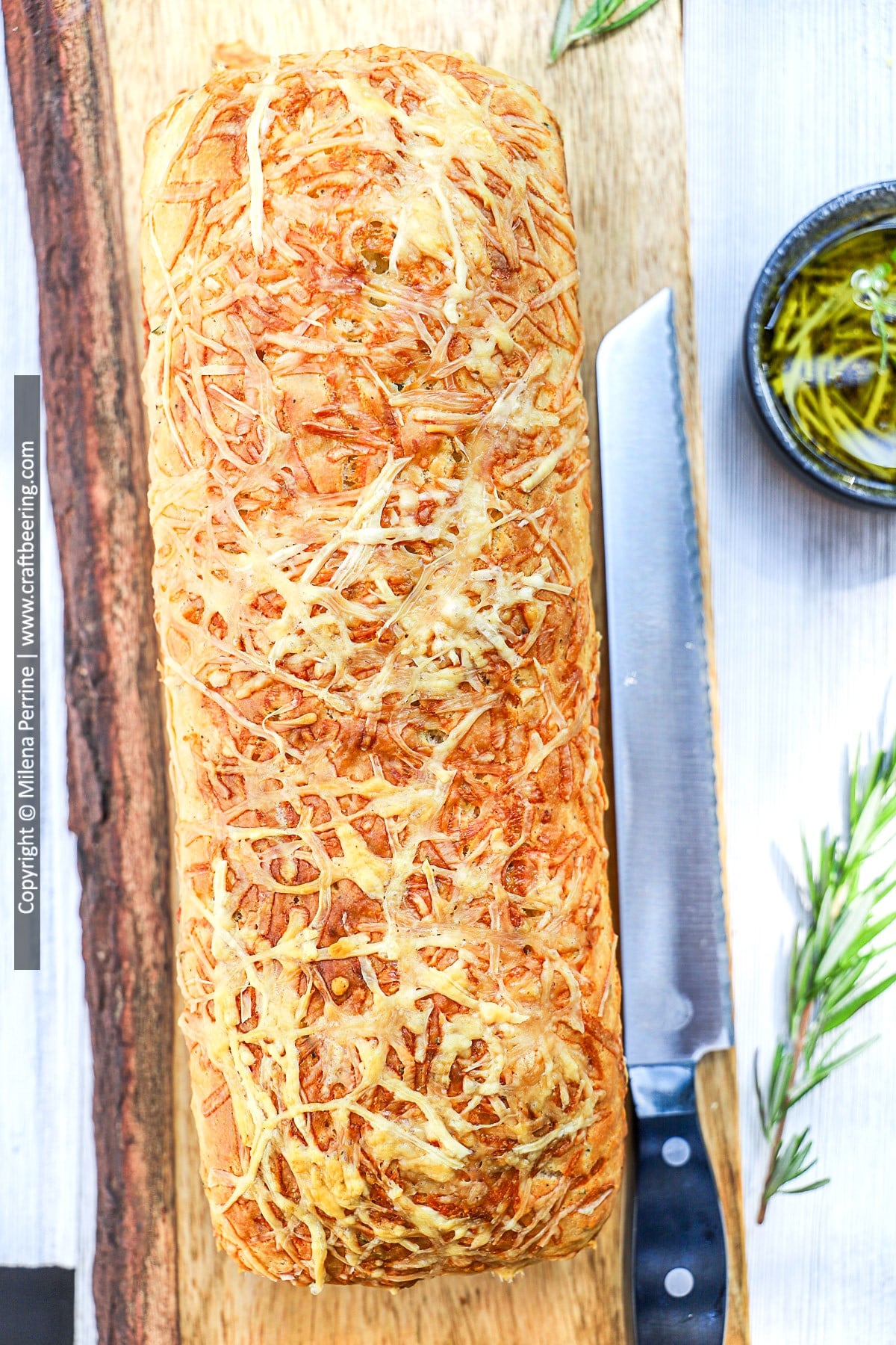 A loaf of rosemary parmesan bread no yeast is shown from above on a cutting board next to a serrated bread knife.