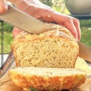 Loaf of rosemary parmesan bread is shown being sliced with a serrated knife over a wood cutting board.