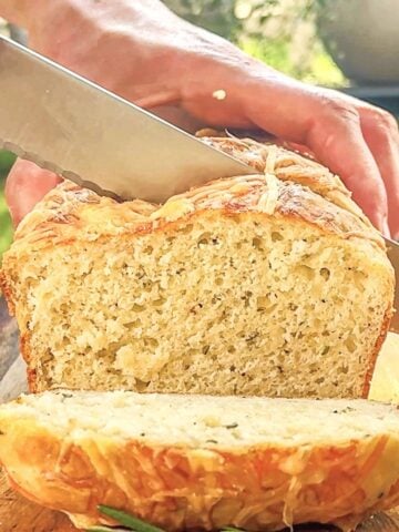 Loaf of rosemary parmesan bread is shown being sliced with a serrated knife over a wood cutting board.