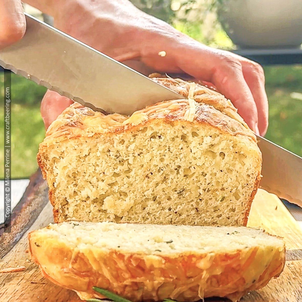 Loaf of rosemary parmesan bread is shown being sliced with a serrated knife over a wood cutting board.