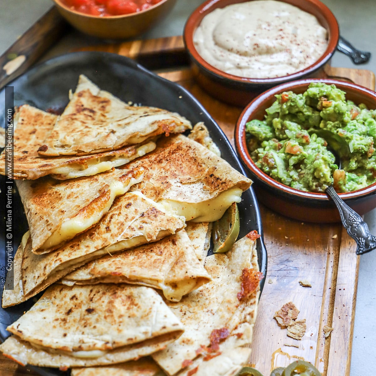 Cheese quesadilla wedges are shown arranged over a black platter with guacamole and quesadilla sauce.