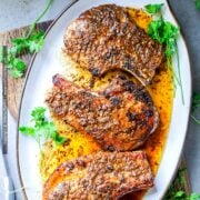 Thick cut pork chops are shown perfectly cooked with a golden brown sear on a white oval platter drizzled with pan sauce and garnished with fresh parsley