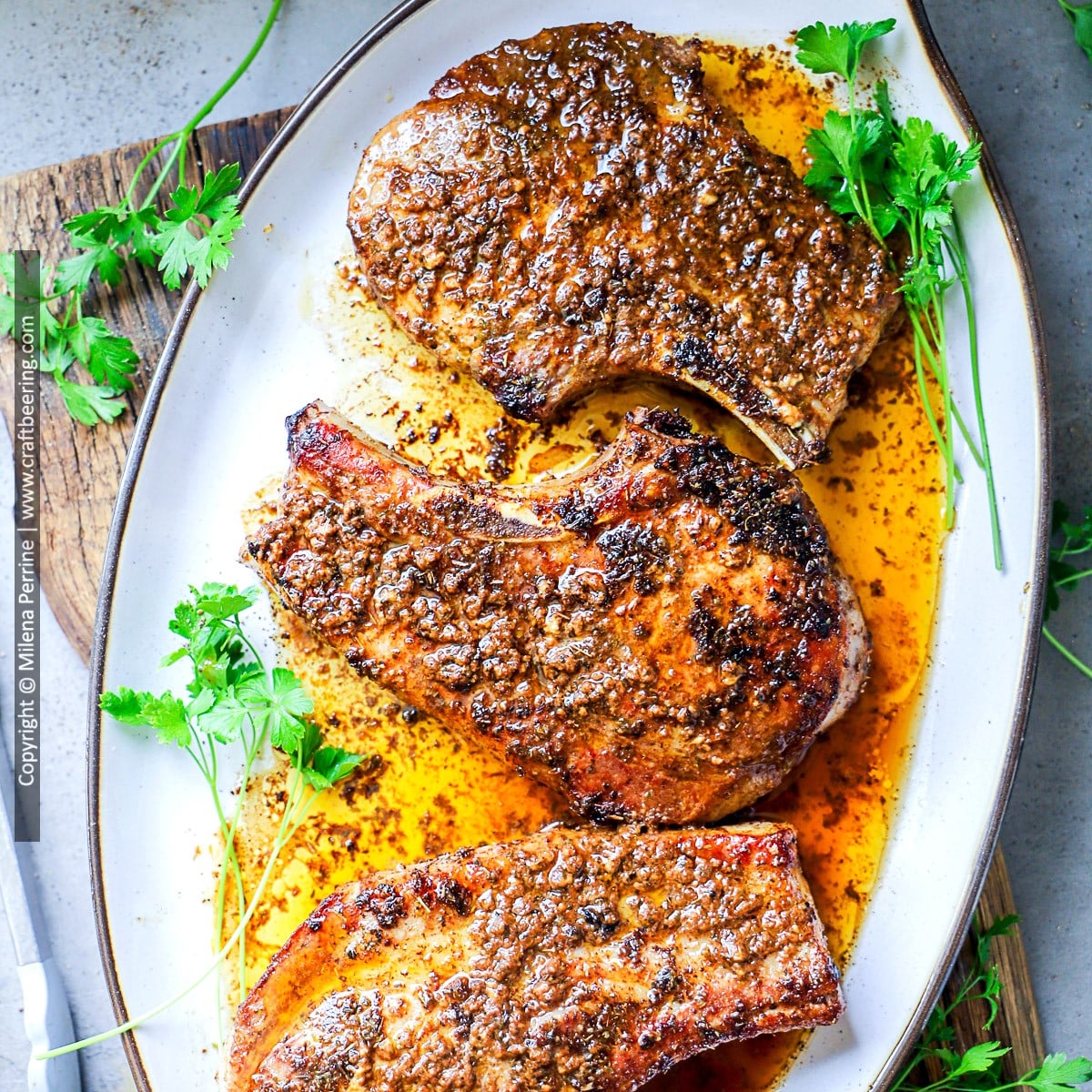 Thick cut pork chops are shown perfectly cooked with a golden brown sear on a white oval platter drizzled with pan sauce and garnished with fresh parsley
