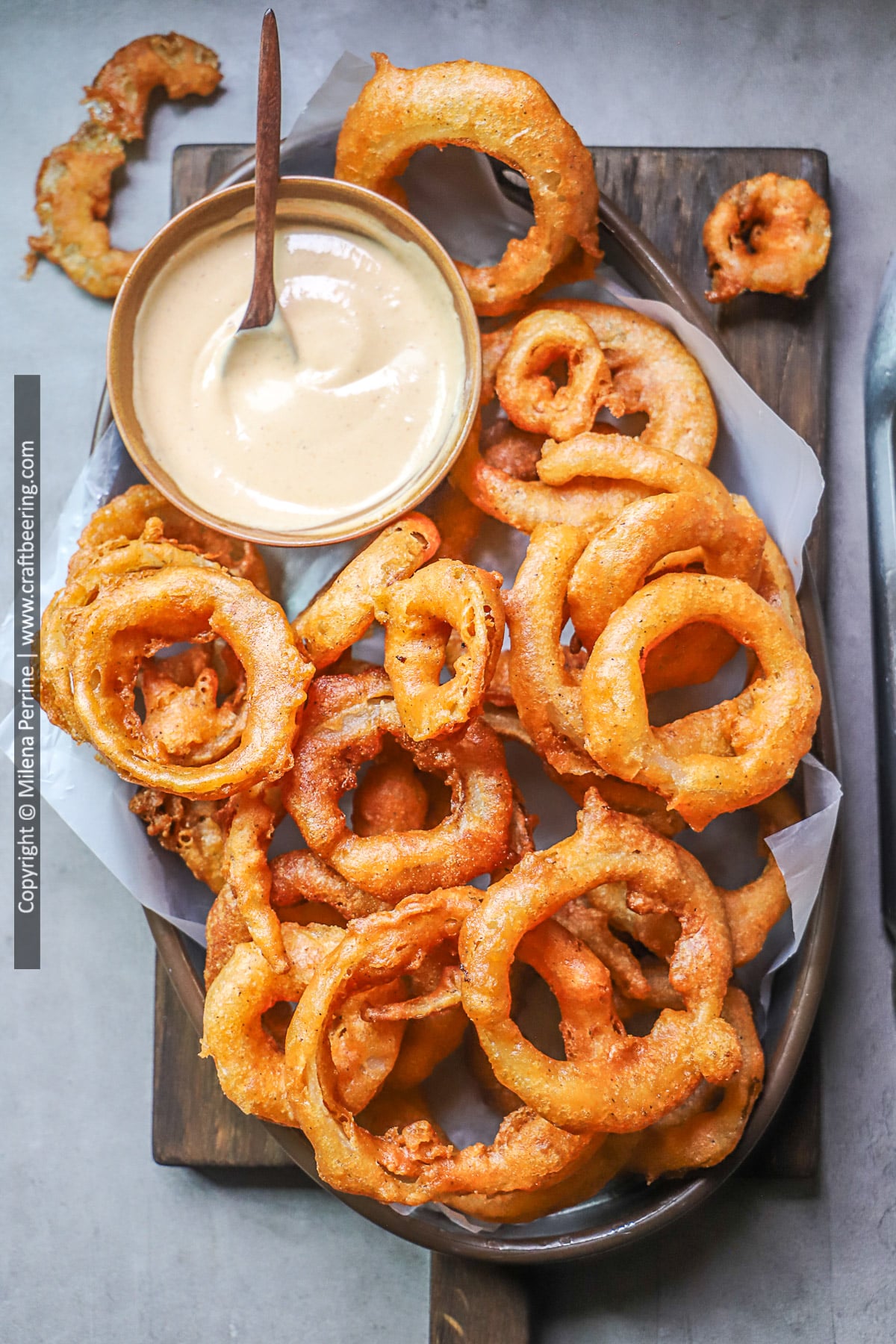 Homemade onion rings are shown piled up in an oval tin dish with an onion ring sauce plated in a round dish.